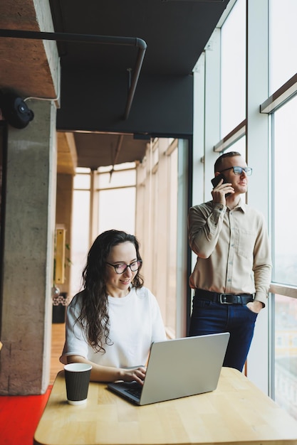 A woman in glasses sits at a table and works on a laptop a male colleague in the background is talking on the phone A working day in a modern office