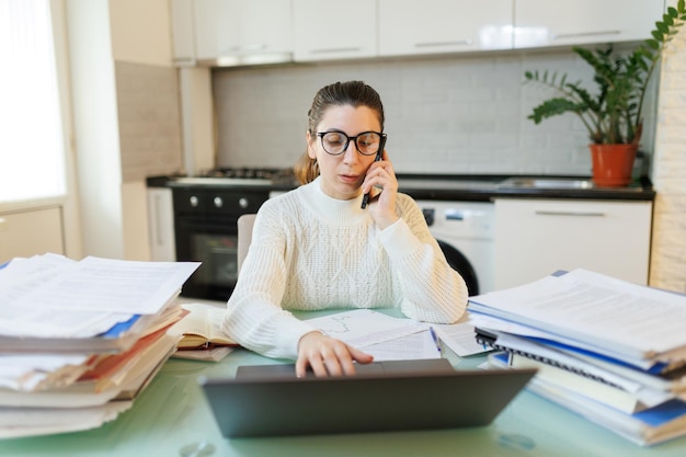 Foto una donna con gli occhiali stanca dal lavoro si siede al tavolo dell'ufficio di casa sua impegnata in una conversazione telefonica