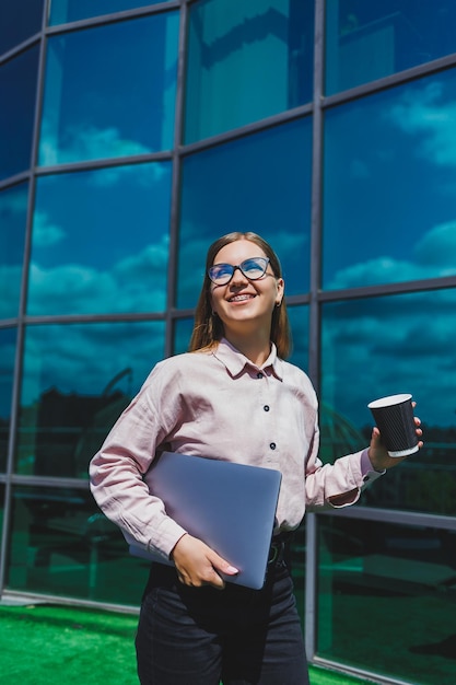 Una donna con gli occhiali con il caffè nelle sue mani contro un grattacielo paesaggio urbano del centro finanziario di edificio moderno imprenditrice di successo con gli occhiali