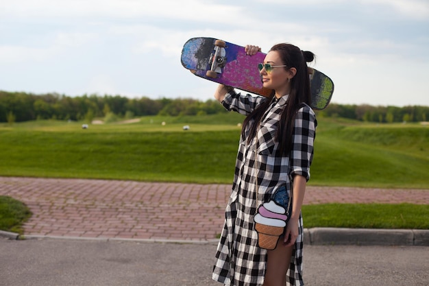 The woman in glasses with cute mini bag is standing with an old skate in her hand on the background