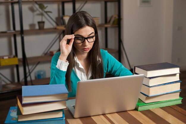 Una donna con gli occhiali con molti libri e un laptop grigio è seduta al tavolo