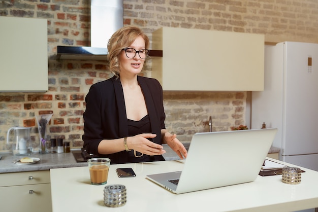 A woman in glasses works remotely on a laptop in her kitchen. A blond girl gesticulating discusses with her business partners on a video conference at home.