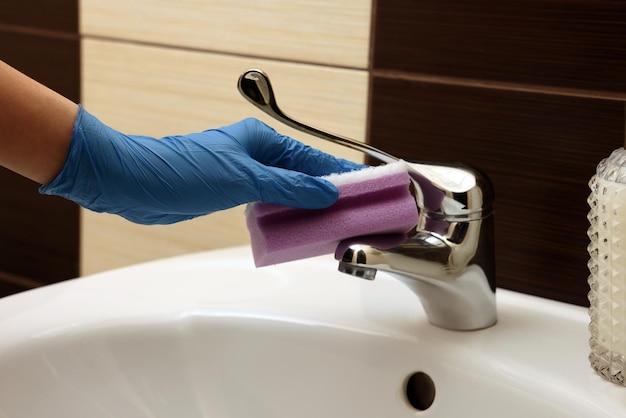 Woman in gloves cleaning faucet of bathroom sink with sponge closeup