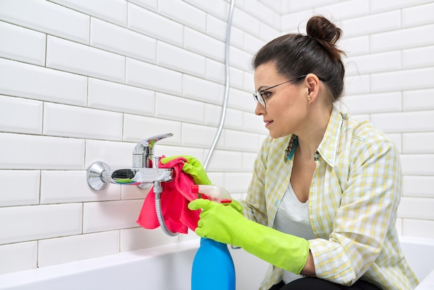 Woman in gloves with detergent and rag doing bathroom cleaning, washing and polishing shower mixer sink, white ceramic tile wall background copy space