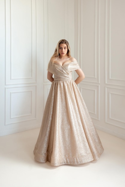 A woman in a gold ball gown stands in a room with a white wall behind her.