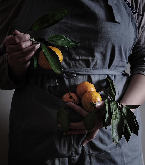 Photo woman in gray apron holds orange tangerines with leaves in her hands.