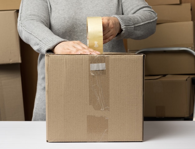 Woman in a gray sweater holds a roll of duct tape and packs brown cardboard boxes on a white table, behind a stack of boxes. moving concept