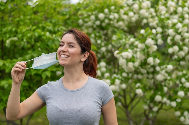 Женщина в серой футболке радостно снимает медицинскую маску по окончании карантина Девушка празднует окончание карантина на прогулке в парке Свободное дыхание без аллергии