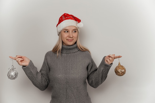 a woman in a gray woolen sweater and a santa hat holds christmas decorations in her hands