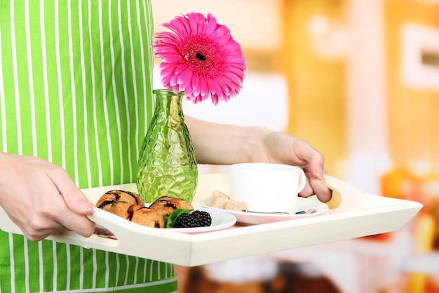 Woman in green apron holding wooden tray with breakfast on bright background