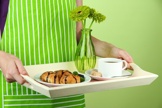 Woman in green apron holding wooden tray with breakfast on color background