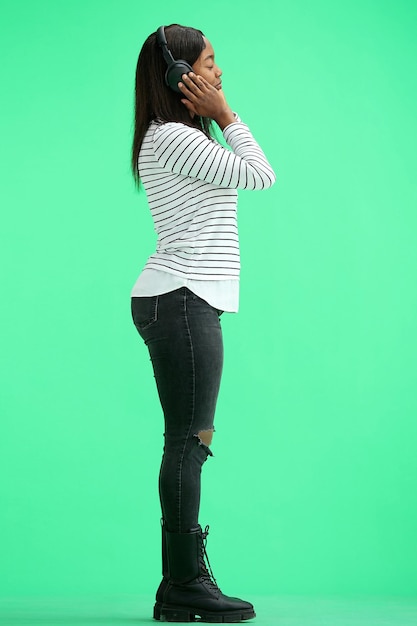 A woman on a green background in full height listening to music with headphones