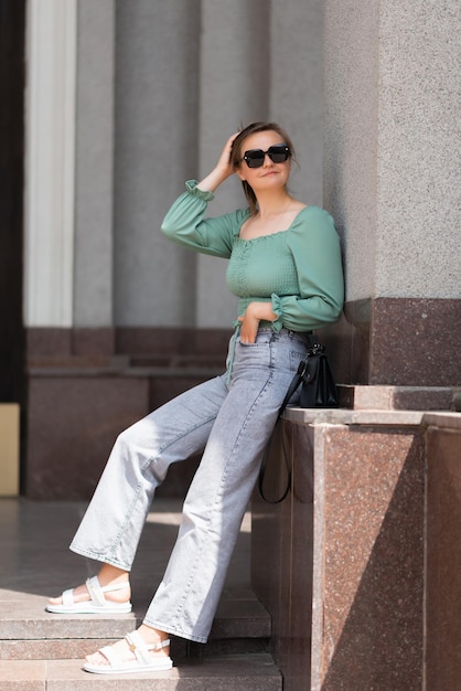 A woman in a green blouse and jeans is standing by the wall