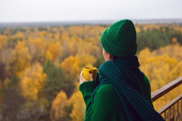 Woman in a green knitted sweater and a hat stands with a yellow mug on the balcony against the background of an autumn forest