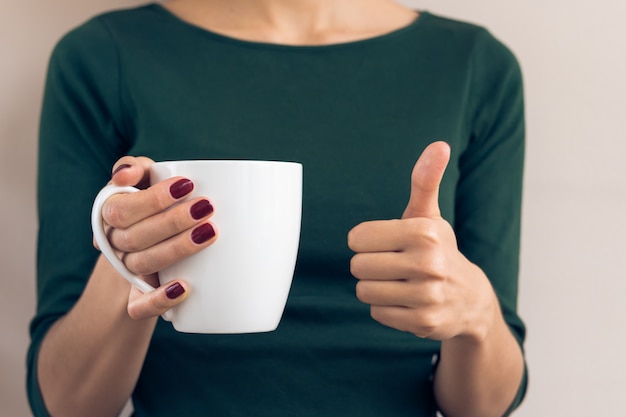 Woman in a green T-shirt and a maroon manicure holding white cup and showing thumbs up
