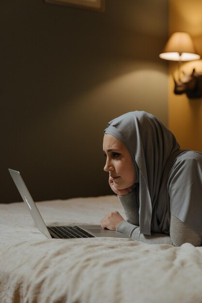 A woman in a grey hoodie is laying on a bed with a laptop