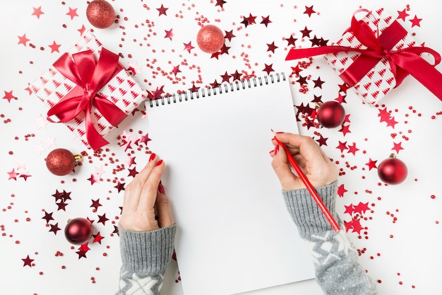 Woman in grey sweater writting checklist of plans and dreams for next year. Wish list for Christmas. To Do List for New 2020 year with red Holiday decor.