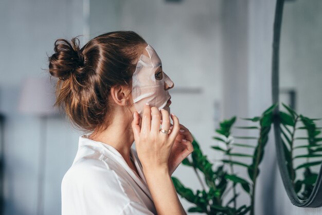 Woman grooming her face in the bathroom with a mask on her face in front of the mirror.