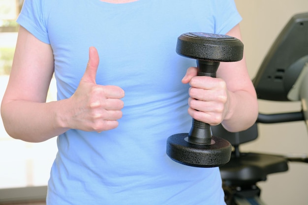 Woman in the gym holds a metal dumbbell and showing a thumbs up