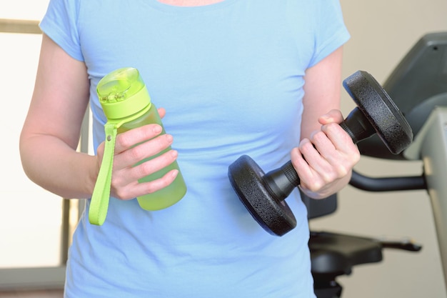 Woman in the gym holds a metal dumbbell and a water bottle in her hands