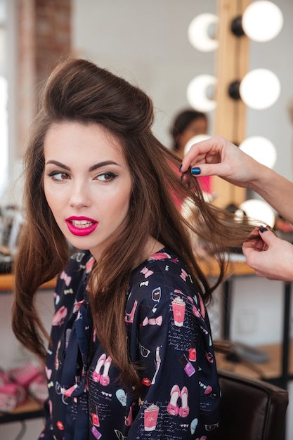 Woman hair stylist making hairdo to attractive young model with bright pink lipstick sitting in beauty salon