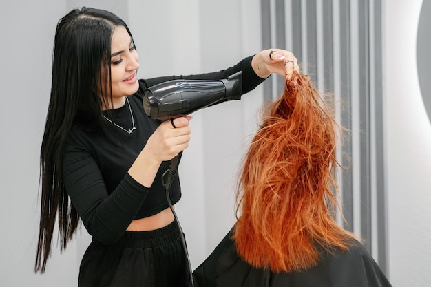 Woman hairdresser does hair styling with hairdryer to client in beauty salon in front of mirror Professional stylist