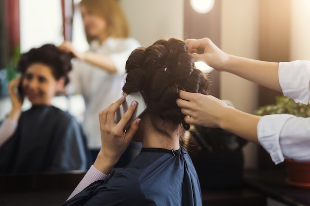 Woman hairdresser making hairstyle for long hair of young female talking on smartphone in beauty salon
