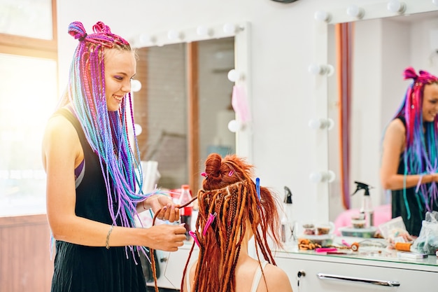 Parrucchiere donna con trecce afro colorate intreccia a ragazza zenzero dreadlocks stile hippie processo di tessitura dei capelli con kanekalon servizi di salone di bellezza luce estiva