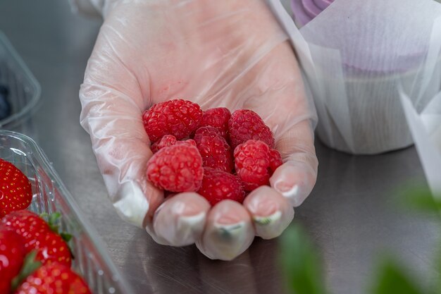 woman hand chef in a glove holds a handful of raspberries to decorate the cake. 