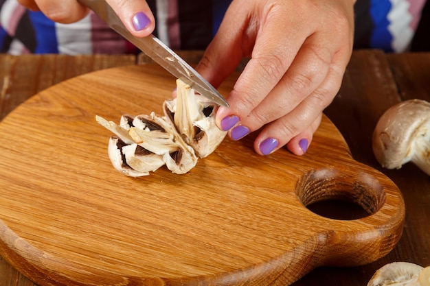 Woman hand cuts champignon mushrooms with a knife on a round wooden board