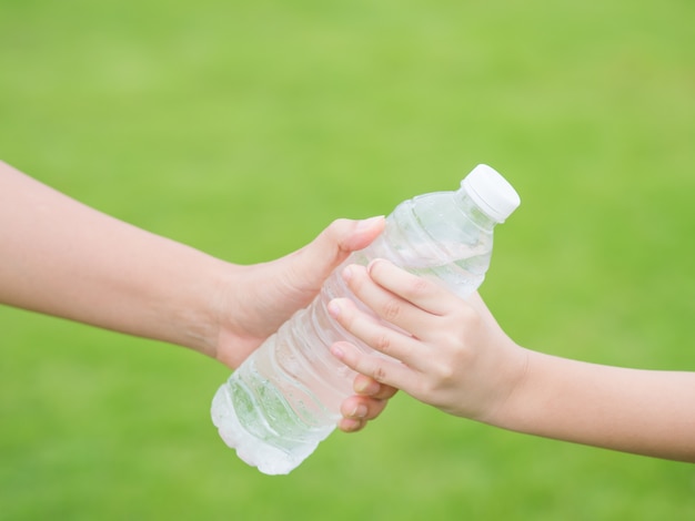 Woman hand giving bottle of fresh water to child on the green grass background