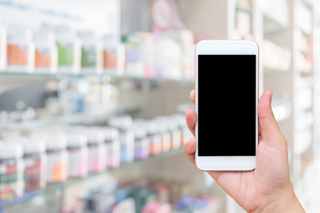 Smartphone mobile dello schermo in bianco della stretta della mano della donna sopra la medicina sugli scaffali del negozio della farmacia sfocatura dello sfondo