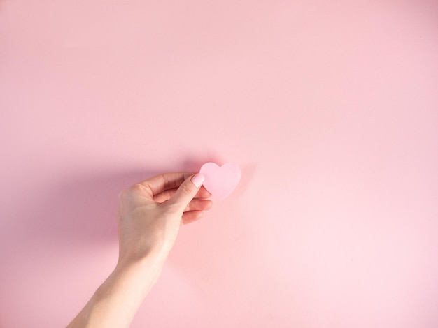 Woman hand hold heart shape pink paper on pink background. Celebrating Valentine's Day