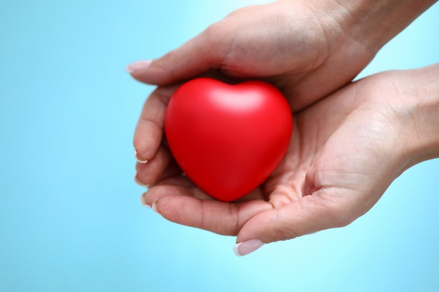 Woman hand hold red toy heart in hand against blue background closeup. Charity people concept