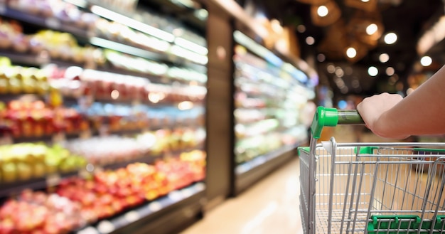 Carrello della spesa del supermercato della stretta della mano della donna con la frutta e la verdura fresche organiche della sfuocatura astratta sugli scaffali nel fondo della luce del bokeh defocused del negozio di alimentari