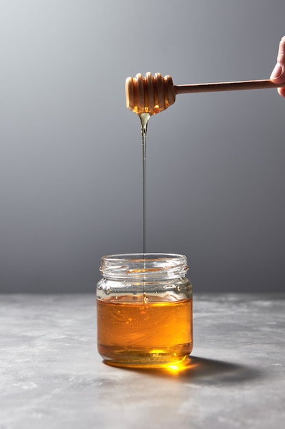 Woman hand hold a wooden stick with dripping sunflower honey in a glass jar on a gray background