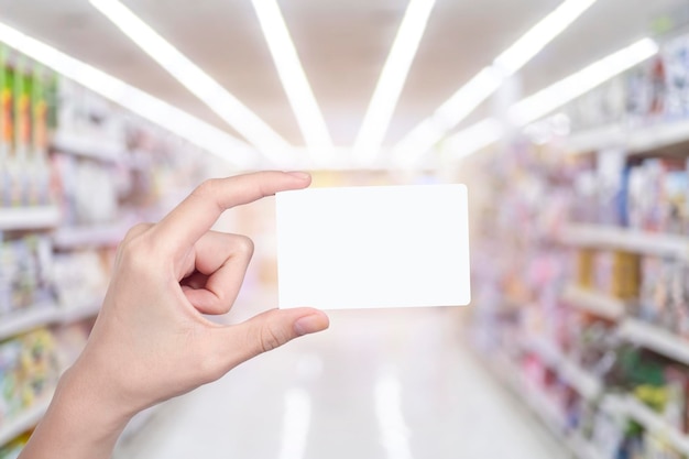 Woman hand holding blank white credit card with blurred abstract background of multicolored cotton clothing on the shelves of fashion shop