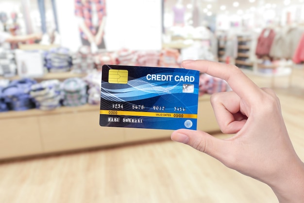 Woman hand holding blue credit card with blurred abstract background of multicolored cotton clothing on the shelves of fashion shop