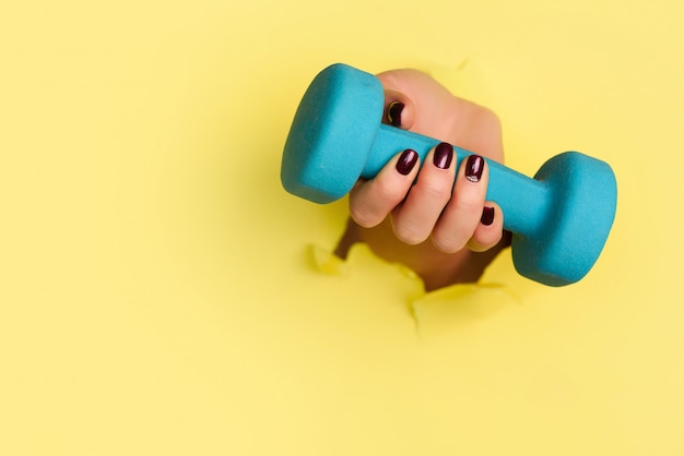 Woman hand holding blue dumbbell on yellow background. 