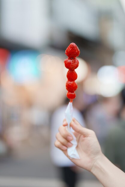 Woman hand holding caramel coated strawberry skewer at night market Street Food and travel concept