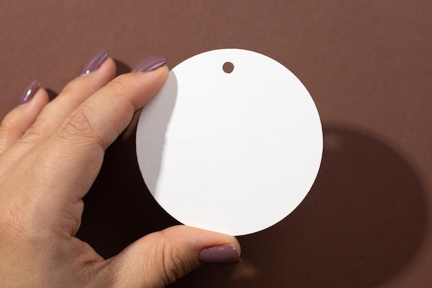 Woman hand holding cardboard white blank tag of round shape with little hole for clothes in upper part in center of brown background Tag mock up Copy space