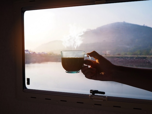 Woman hand holding cup of hot coffee in the morning over glass window with mountian view background.