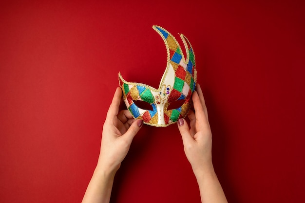 Woman hand holding  festive, colorful mardi gras or carnival mask over red wall.Flat lay, top view