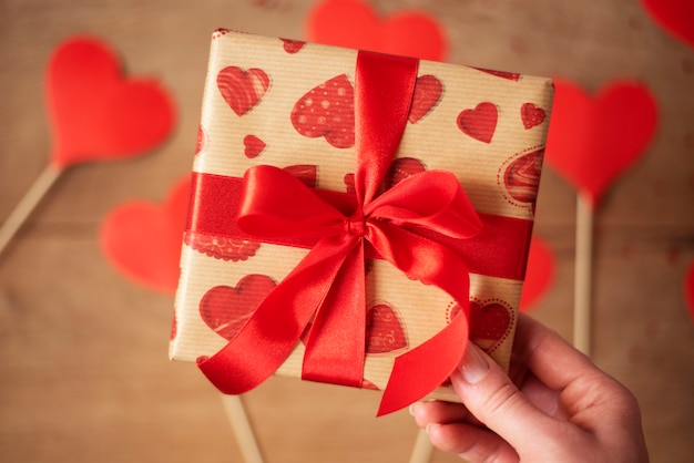 Woman hand holding gift box with red bow. Many hearts on a wooden  in defocus. Valentine's Day concept