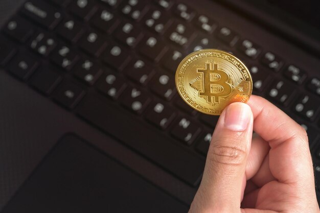 黒いノートパソコンのキーボードの上に金色のビットコインコインを持つ女性の手