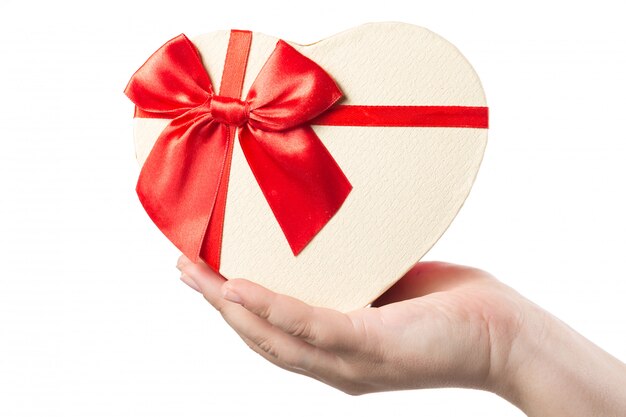 Woman hand holding heart shaped present box with red ribbon isolated on white .
