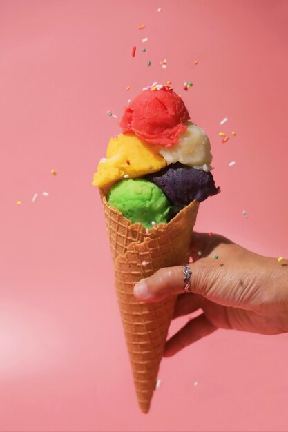 Woman Hand holding homemade ice cream cone over pink background with colorful sprinkles in the air.