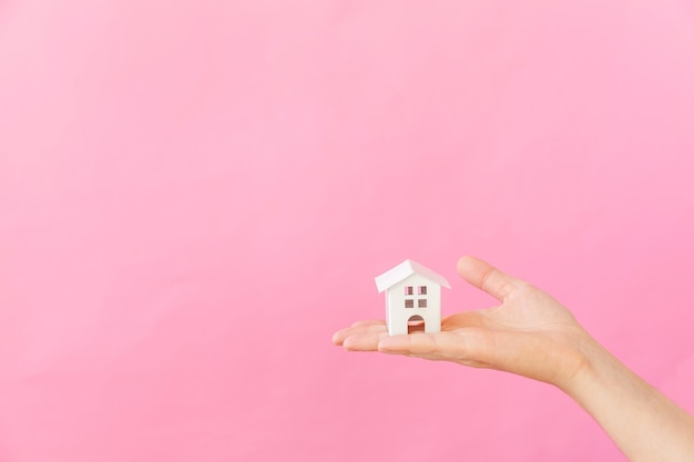 ピンクの背景で隔離のミニチュア白いおもちゃの家を持っている女性の手