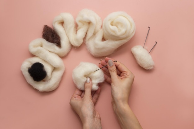 Woman hand holding needle felting kit felted woolen needle and skein of wool on pink table flat lay