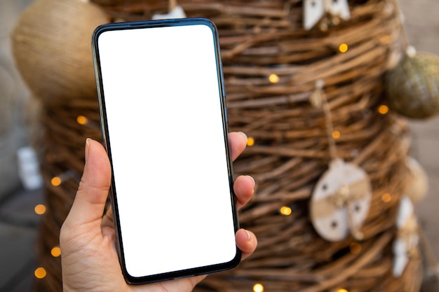 背景に白い画面のクリスマス ツリーと携帯電話を持つ女性の手
